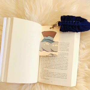 Op de afbeelding: Een open boek met een kleurrijke bladwijzer en een marineblauwe macramé kwast. De bladwijzer toont een illustratie van een wolk en een bruin object. De kwast is bevestigd met een goudkleurige sluiting.