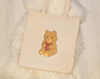 May include: A beige tote bag featuring a cartoon bear holding a red heart. The bear is brown with a yellow outline. The bag has a simple design with two handles.