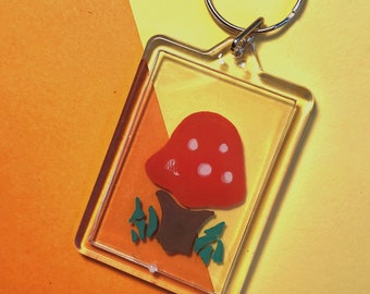 Puede incluir: Llavero acrílico transparente con un hongo rojo con puntos blancos, tallo marrón y hojas verdes. El llavero tiene un anillo de metal plateado. El fondo es una división diagonal de naranja y amarillo.