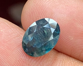 Puede incluir: Una piedra preciosa de corte ovalado con una paleta de colores azul verdoso profundo. La piedra presenta una superficie facetada que refleja la luz, mostrando variaciones internas de tono y claridad en toda la estructura cristalina de la gema.