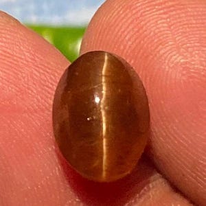 Puede incluir: Una piedra preciosa ovalada de color marrón con un efecto brillante e iridiscente. La piedra tiene un color marrón claro con un sutil brillo dorado.