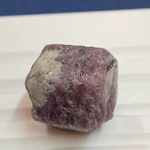 May include: A natural, unpolished gemstone in a deep pink colour, with a rough, textured surface. The stone has patches of white and grey, with small dark green inclusions. The overall shape is a roughly rectangular cube.