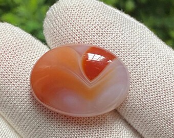 May include: An oval-shaped gemstone with a polished surface. The stone features a blend of orange, red, and white hues, with a prominent heart-shaped pattern in the center. The gemstone is held against a neutral-colored fabric background.