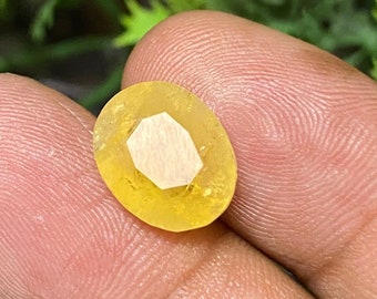 May include: A yellow oval-shaped gemstone with a faceted cut. The gemstone is held in a hand.