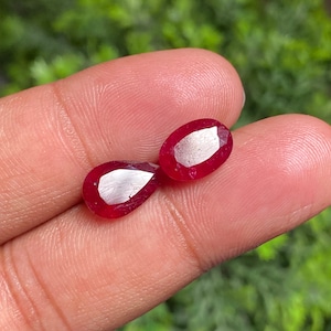 Op de afbeelding: Twee peervormige rode edelstenen, waarschijnlijk robijnen, worden in een hand gehouden. De edelstenen zijn glad en gepolijst, met een lichte glans.