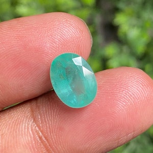 May include: A vibrant green oval-shaped gemstone with a faceted cut. The gemstone is held in a hand, showcasing its colour and clarity.