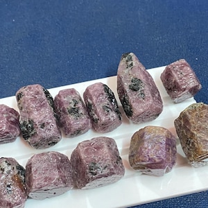 May include: A collection of raw ruby gemstones, displaying a range of shapes and sizes. The rubies exhibit a deep pinkish-red hue with dark inclusions, resting on a white surface against a blue backdrop. These natural stones are uncut and unpolished.
