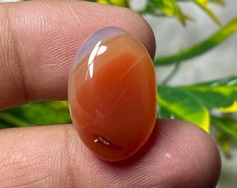 May include: An oval-shaped gemstone with a polished surface. The stone features a gradient of colors, transitioning from a pale lavender hue at the top to a vibrant orange-red at the base. The gemstone is held between fingers, with a blurred green and yellow background.