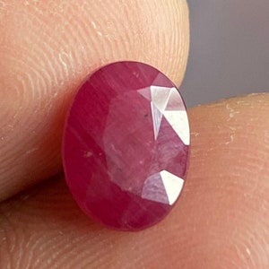 May include: A vibrant red oval-shaped gemstone with a faceted cut. The gemstone is set against a neutral background.