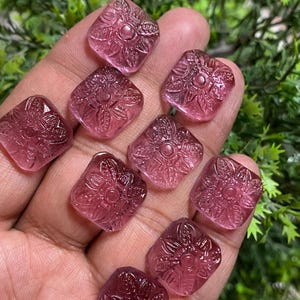 Puede incluir: Diez cabujones cuadrados de color rosa con un diseño floral tallado. Los cabujones son translúcidos y tienen un ligero brillo.