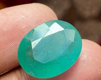 May include: A large, oval-shaped, faceted emerald gemstone with a green hue. The gemstone is set in a clear, faceted setting.