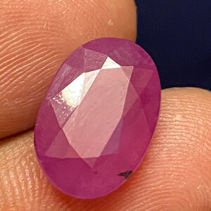 Puede incluir: Una gema ovalada de color rosa vibrante con un corte facetado. La gema tiene un ligero tono púrpura y está engastada en un engaste claro y facetado.
