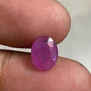 Puede incluir: Una gema de rubí ovalada y facetada. La gema es de un color rojo rosado vibrante, con inclusiones internas visibles. Las facetas captan la luz, creando un efecto brillante. El rubí se sostiene entre dos dedos.