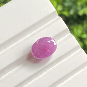 May include: A faceted, oval-shaped, pink gemstone. The gem is set against a white, ridged surface, with a blurred green background. The stone's color is a vibrant, medium pink, with visible internal structures.