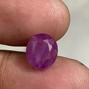 以下が含まれることがあります： ファセット加工された楕円形のルビーの宝石。宝石は、光を捉える複数のファセットを持つ、深く飽和したピンクがかった赤色です。石は2本の指の間に保持され、そのサイズと形状を示しています。