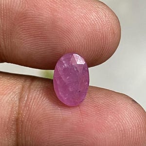 Può includere: Una gemma rosa sfaccettata, di forma ovale. La gemma è tenuta tra due dita, mostrando le sue superfici lucide e la sua chiarezza interna. Il colore della pietra varia dal rosa chiaro al medio, con inclusioni interne visibili.