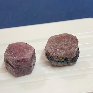 Puede incluir: Dos gemas en bruto de color púrpura rojizo con una estructura cristalina. Una piedra tiene una banda oscura en la base. Las gemas se exhiben sobre una superficie blanca, con un fondo azul.