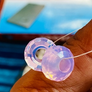 May include: Two iridescent purple glass beads with a faceted design. The beads are strung on a white thread.