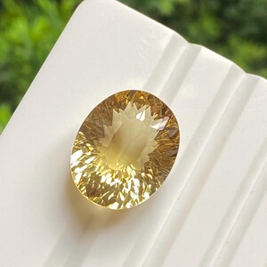 Op de afbeelding: Een gefacetteerde, ovaalvormige edelsteen met een warme, goudgele tint. De edelsteen ligt op een wit oppervlak, met een vage groene achtergrond. De slijping van de steen creëert een steruitbarsting-effect.