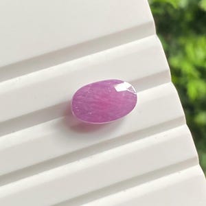 May include: A faceted, oval-shaped, pink gemstone. The gem has a polished surface with multiple facets that reflect light. The stone is set against a white, grooved surface, with a blurred green background.