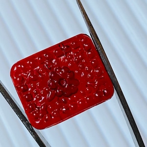 May include: A red square-shaped glass tile with a raised floral design. The tile is being held by two silver tweezers.