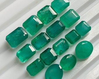 May include: A collection of 13 emerald green gemstones of various sizes and shapes, including round and rectangular cuts. The gemstones are arranged on a white surface.