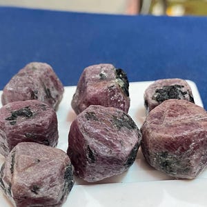 May include: A collection of rough-cut ruby gemstones, each displaying a deep pinkish-red hue with dark inclusions. The stones are presented on a white surface, showcasing their natural, unpolished texture. These gemstones are ideal for jewelry making or collecting.