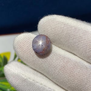 May include: A small, spherical gemstone, displaying a blend of purple and reddish-brown tones. The stone is held in a white glove, highlighting its smooth texture and natural colour variations. The gemstone appears to be a bead or a small decorative element.