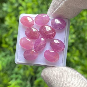 May include: A collection of ten oval-shaped, pink gemstones displayed in a white plastic container. The gemstones vary in shades of pink, with some appearing more translucent than others. The background is a blurred green.