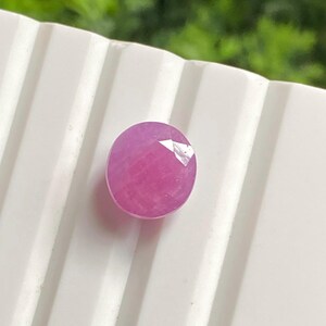 May include: A round, faceted, pink gemstone. The gem is a vibrant magenta color and sits on a white, ridged surface. The background is a blurred green, suggesting a natural setting. This gemstone is suitable for jewelry making.