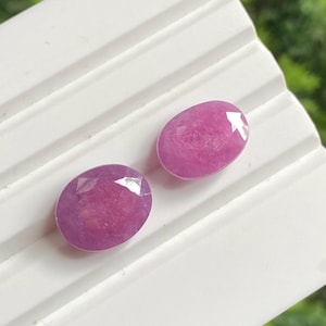 May include: Two oval-shaped, faceted, pink gemstones. The gems are set against a white, ridged surface, with a blurred green background. The gemstones have a polished, reflective surface, showcasing their internal structure and colour.