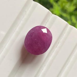 Peut inclure: Une pierre précieuse rubis ovale et facettée. La gemme est d'un rouge-rose profond et a une surface polie et réfléchissante. Le rubis est posé sur un fond blanc strié, avec un feuillage vert visible en arrière-plan.