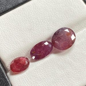 May include: Three oval-shaped gemstones in a jewelry box. The gemstones are red with a pink hue and have a faceted cut. The gemstones are arranged in order of size, with the largest gemstone at the top and the smallest gemstone at the bottom.