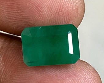 May include: A vibrant green emerald gemstone, cut into a rectangular shape with beveled edges. The emerald is set in a clear, faceted setting.