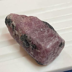 May include: A raw ruby gemstone, displaying a deep pink colour with dark grey inclusions. The natural stone has a textured surface and an irregular shape, highlighting its unpolished state. The ruby is presented on a white surface.