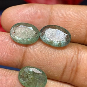 May include: Three oval-shaped, faceted gemstones with a translucent, pale green hue. Each stone features a polished surface with visible internal inclusions and natural textures, displaying a mix of light and dark green tones throughout the structure.