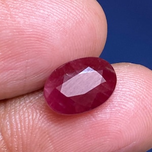 Pode incluir: Uma pedra preciosa de rubi facetada em forma oval. O rubi é de cor vermelha profunda com múltiplas facetas que refletem a luz. A pedra preciosa é segurada entre dois dedos contra um fundo azul.
