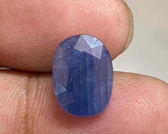 May include: A faceted, oval-shaped, deep blue gemstone. The gem has a polished surface with multiple facets that catch the light, creating a sparkling effect. The stone is held between fingers, showcasing its color and clarity.