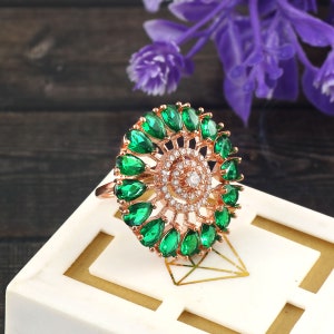 May include: A rose gold ring with a large green gemstone center surrounded by smaller green gemstones. The ring is on a white surface.