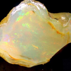 Könnte beinhalten: Ein roher, weißer Opal-Edelstein mit irisierenden Schimmern in Grün, Blau und Orange. Der Opal ist in eine Matrix aus braunem und gelbem Gestein eingebettet.