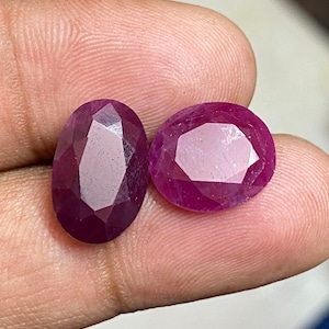 May include: Two oval-shaped, faceted ruby gemstones with a deep pink hue. The gemstones are set against a light pink background.