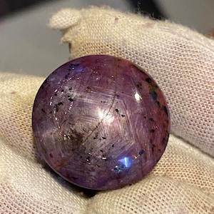 Può includere: Una pietra preziosa rotonda di colore viola con un motivo a forma di stella e inclusioni nere.