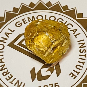 Könnte beinhalten: Ein gelber Edelstein mit einer rauen, facettierten Oberfläche. Der Edelstein liegt auf einem weißen Etikett mit dem Text "International Gemological Institute IGI 1975".