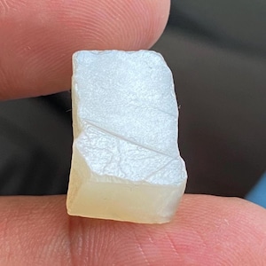 May include: A small, rectangular, off-white gemstone with a slightly translucent appearance. The stone has a textured surface with subtle lines and a pale yellow base. The edges are uneven, suggesting a natural, unpolished form.