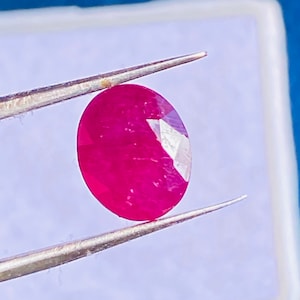 Puede incluir: Una gema de rubí de corte ovalado de color rojo intenso con una superficie facetada, sostenida firmemente por un par de pinzas de metal plateado. La gema muestra reflejos de luz internos y claridad, destacando su estructura pulida y facetada.