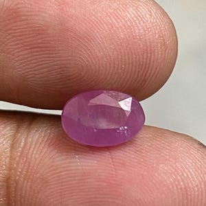 以下が含まれることがあります： 楕円形にカットされた、ピンク色のファセット宝石。 宝石は2本の指の間に挟まれ、研磨された表面と透明度を示しています。 石の色は、薄いピンクから中程度のピンクまであり、光を捉えるファセットが見えます。