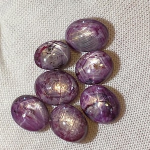 May include: Seven oval-shaped, star ruby gemstones in varying shades of purple and pink. The gemstones have a polished, reflective surface with a star-like effect. They are displayed on a white, textured fabric.
