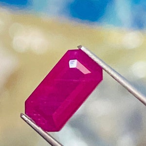以下が含まれることがあります： ピンセットで保持された長方形にカットされた濃い赤色の宝石
