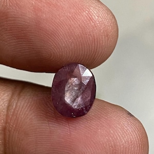 Peut inclure: Une pierre précieuse de type rubis, de forme ovale et facettée. La gemme est d'un rouge-violet profond, avec des inclusions internes visibles. La pierre est tenue entre deux doigts, mettant en valeur ses facettes polies et sa beauté naturelle. Cette pierre convient à la joaillerie.