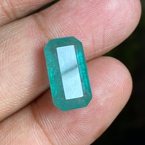 Puede incluir: Una gema de esmeralda verde vibrante con un corte rectangular y un acabado pulido. La gema está engastada en un engaste rectangular transparente.